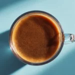 Top-view square image of a clear mug filled with smooth homemade coffee on a bright, minimal background.